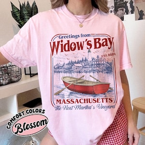 May include: A light pink t-shirt featuring a graphic print with the text Greetings from Widow's Bay, EST. 1681, MASSACHUSETTS, and The Next Martha's Vineyard. The design includes a red rowboat on water, a lighthouse, and coastal buildings.