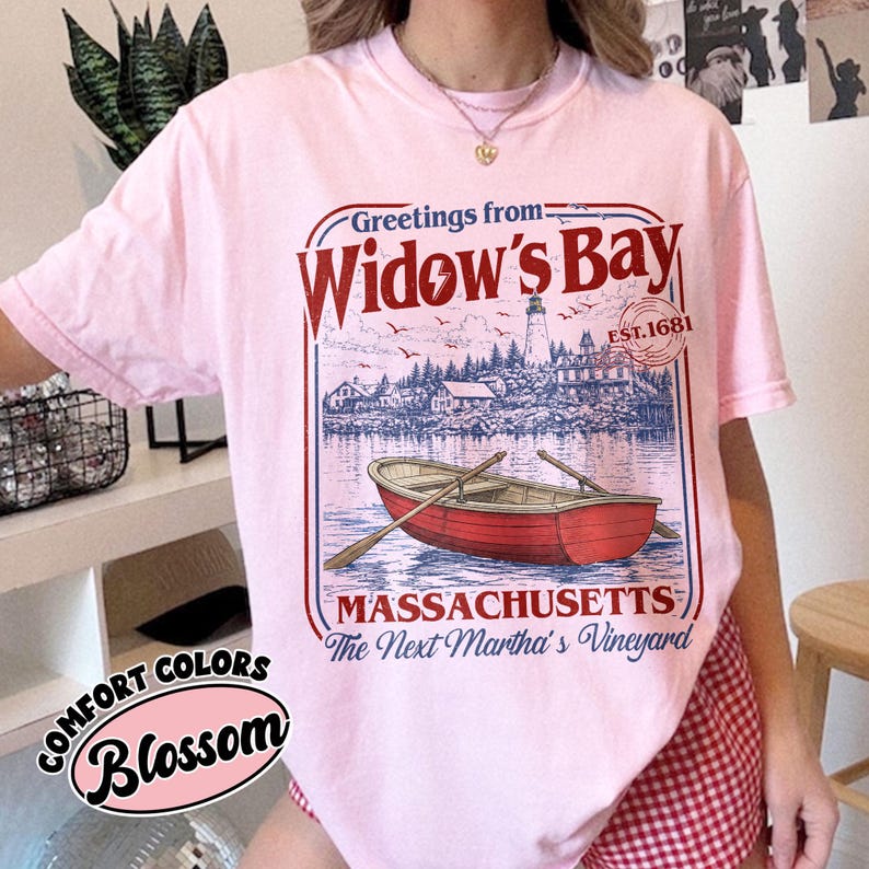 May include: A light pink t-shirt featuring a graphic print with the text Greetings from Widow's Bay, EST. 1681, MASSACHUSETTS, and The Next Martha's Vineyard. The design includes a red rowboat on water, a lighthouse, and coastal buildings.