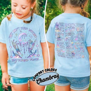 May include: Light blue t-shirt with the text "THEY DIDN'T BURN WITCHES THEY BURNED WOMEN" and a graphic of a crescent moon, skeleton hands, and stars. The back of the shirt has a doodle-style design. The shirt is labeled "Comfort Colors Chambray."