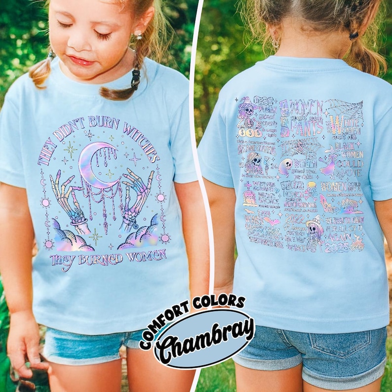 May include: Light blue t-shirt with the text "THEY DIDN'T BURN WITCHES THEY BURNED WOMEN" and a graphic of a crescent moon, skeleton hands, and stars. The back of the shirt has a doodle-style design. The shirt is labeled "Comfort Colors Chambray."