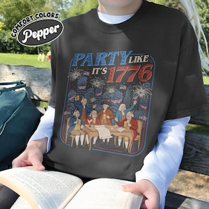 Puede incluir: Camiseta gris oscuro con la frase "PARTY LIKE IT'S 1776". El dise&ntilde;o gr&aacute;fico presenta una ilustraci&oacute;n vintage de personas celebrando con fuegos artificiales y banderas. La camiseta se lleva sobre una camiseta blanca de manga larga.