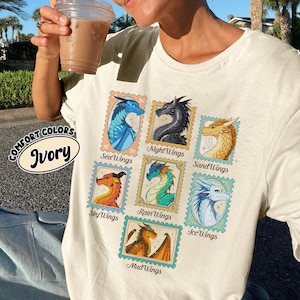 May include: Ivory-colored t-shirt featuring colorful dragon illustrations within postage stamp frames. The shirt displays various dragon types, including SeaWings, NightWings, SandWings, SkyWings, RainWings, IceWings, and MudWings. The text "COMFORT COLORS" and "Ivory" are also visible.