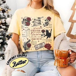 Puede incluir: Una camiseta amarilla con un estampado gr&aacute;fico de rosas rojas, un gato negro, una pistola y texto que dice "Let me know which stars you prefer the ones above you or the ones I make you see". El texto "House of Mirrors" tambi&eacute;n es visible en la camiseta.