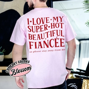 May include: A light pink t-shirt with the text "I LOVE MY SUPER HOT BEAUTIFUL FIANC&Eacute;E so please stay away from me" in red. The shirt also has a "Comfort Colors Blossom" logo.