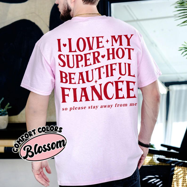May include: A light pink t-shirt with the text "I LOVE MY SUPER HOT BEAUTIFUL FIANC&Eacute;E so please stay away from me" in red. The shirt also has a "Comfort Colors Blossom" logo.