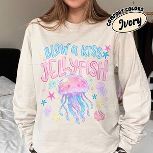 May include: Ivory long-sleeve shirt with a colorful jellyfish graphic. The design includes the text "BLOW A KISS JELLYFISH" in blue and pink, along with starfish and seashell illustrations. The shirt is labeled "COMFORT COLORS" and "Ivory".