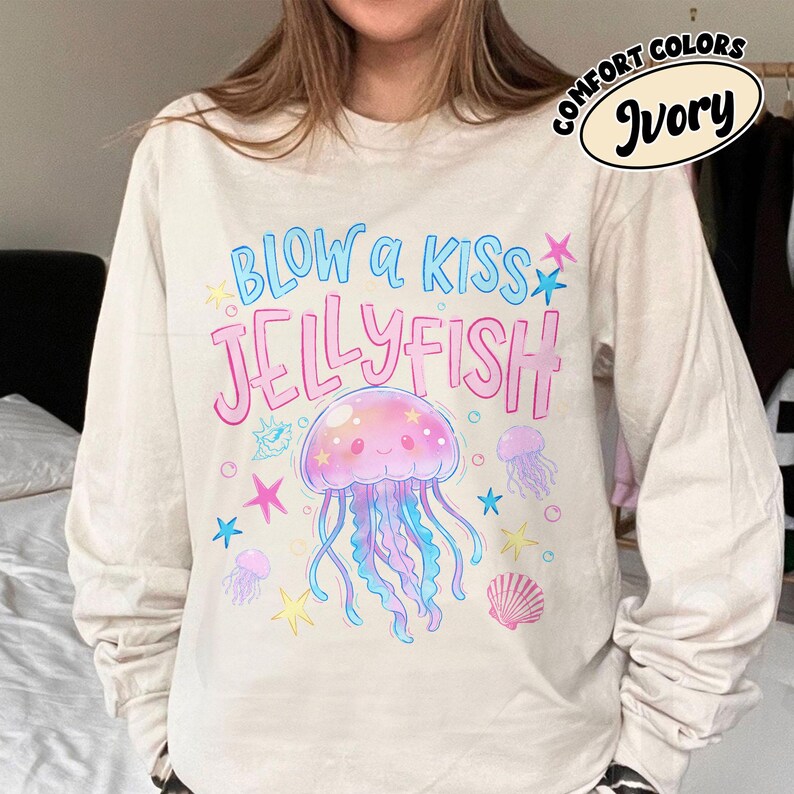 May include: Ivory long-sleeve shirt with a colorful jellyfish graphic. The design includes the text "BLOW A KISS JELLYFISH" in blue and pink, along with starfish and seashell illustrations. The shirt is labeled "COMFORT COLORS" and "Ivory".