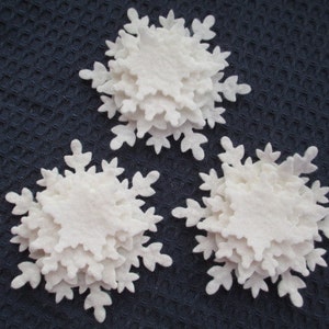 May include: Three white felt snowflakes, each made of multiple layers, are arranged on a dark blue textured surface.