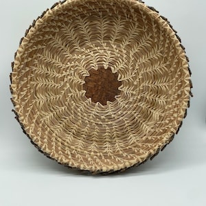 Puede incluir: Una cesta redonda y artesanal con una estética natural y terrosa. La cesta presenta un diseño tejido en tonos beige y marrón, con un borde marrón oscuro similar a la corteza. El centro de la cesta tiene un parche más oscuro y texturizado.