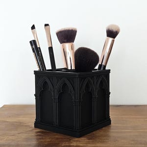 May include: Black makeup brush holder with an arched design, containing various makeup brushes. The brushes have black handles and soft bristles. The holder sits on a wooden surface.