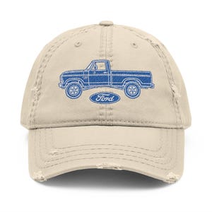 May include: A distressed beige baseball cap featuring a blue graphic of a vintage Ford pickup truck. The word "Ford" is printed in blue below the truck image. The cap has a pre-worn look with distressed edges.