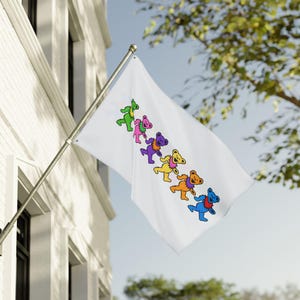May include: A white flag with a row of colorful cartoon bears in green, pink, purple, yellow, orange, and blue. The bears are marching diagonally across the flag. The flag is mounted on a silver pole and is displayed outside a white building.