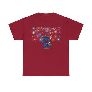 May include: A maroon short-sleeve t-shirt featuring a graphic of a blue bear wearing a tricorn hat and playing a drum. The background displays colorful fireworks, multiple circular skull icons, and a heart-shaped American flag design.