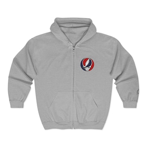 May include: A light gray zip-up hoodie with a front pocket and a hood. The hoodie features a colorful emblem on the chest with a skull and lightning bolt design. The sleeves have a small graphic on the cuff.