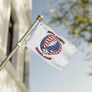 May include: A white rectangular flag attached to a metal pole featuring a central graphic of a skull with a lightning bolt design. The skull is colored with red and white stripes and blue stars. The text "MAKE AMERICA GRATEFUL AGAIN" is printed.