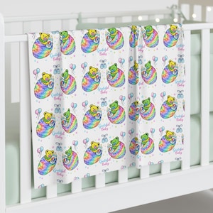 May include: A white baby blanket with a repeating pattern of colorful teddy bears wrapped in rainbow-colored swaddles. The blanket also features the text "Grateful Baby", balloons, and baby shoes.