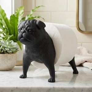 May include: A matte black, pug-shaped toilet paper holder standing on four legs. The sculpture features a detailed canine face and body, designed to support a standard roll of white toilet paper between its front and back sections.