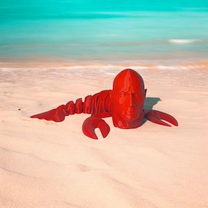 May include: A red sculpture of a lobster with a human face, resting on a sandy beach. The sculpture has a detailed face and lobster claws. The background features a turquoise ocean and sky.