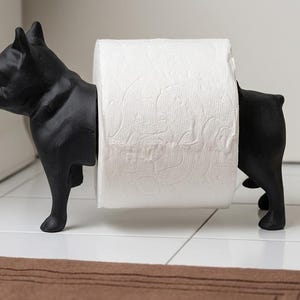 May include: A matte black French bulldog figurine designed as a toilet paper holder. The dog is standing in profile, with its body split into two sections to support a standard roll of white toilet paper between its front and back halves.