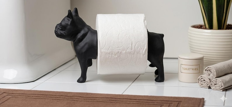 May include: A matte black French bulldog figurine designed as a toilet paper holder. The dog is standing in profile, with its body split into two sections to support a standard roll of white toilet paper between its front and back halves.