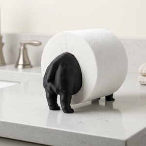 May include: A matte black animal-shaped toilet paper holder featuring a stylized rear end with a tail, supporting a single roll of white toilet paper. The holder stands on four small feet, designed to hold the roll horizontally on a flat surface.