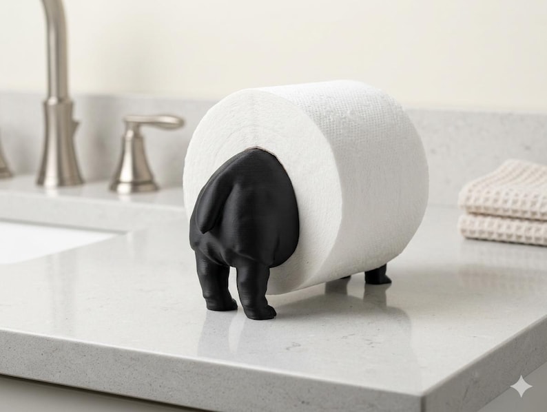 May include: A matte black animal-shaped toilet paper holder featuring a stylized rear end with a tail, supporting a single roll of white toilet paper. The holder stands on four small feet, designed to hold the roll horizontally on a flat surface.
