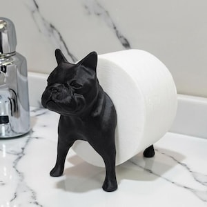 May include: A black French bulldog figurine designed to hold a roll of white toilet paper. The dog stands on four legs with its body positioned to support the paper roll horizontally between its front and back sections in a bathroom setting.
