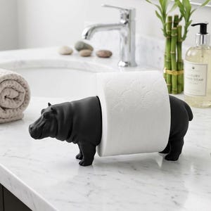 May include: A matte black hippopotamus-shaped toilet paper holder. The sculpture features a realistic hippo form with a smooth finish, designed to hold a standard roll of white toilet paper around its midsection, standing on four sturdy legs.