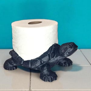 May include: A black turtle-shaped toilet paper holder. The holder is made of a dark material and has a roll of white toilet paper resting on its back. The holder is on a tiled surface with a teal background.