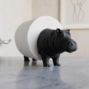 May include: A matte black hippopotamus figurine designed to hold a roll of white toilet paper. The hippo stands on four legs with the roll of paper encircling its midsection, creating a functional holder for bathroom tissue storage.