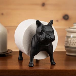 May include: A matte black French bulldog figurine designed as a toilet paper holder. The dog stands on four legs with a cylindrical body shape that supports a standard roll of white toilet paper positioned between its front and back legs.