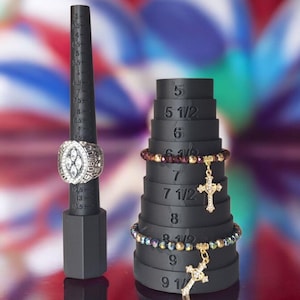 May include: A black ring sizer with a championship ring and a tiered bracelet sizer with bracelets. The ring sizer is marked with sizes, and the bracelet sizer has sizes from 5 to 9 1/2. The bracelets have cross charms.