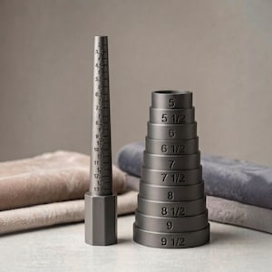 May include: A dark gray metal ring mandrel with a tapered shape and printed size markings from 3 to 13.5, positioned next to a stack of nine dark gray metal ring sizers with engraved size labels ranging from 5 to 9.5 in half-size increments.