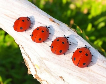 May include: Four red ladybug figurines with black spots and antennae are arranged on a weathered, light-colored wooden branch. The ladybugs are in a row, with a blurred green background.
