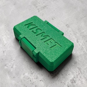 May include: A bright green rectangular box with the word "KISMET" embossed on the top. The box has a textured surface and a hinged design with two clasps. The box is sitting on a gray surface.