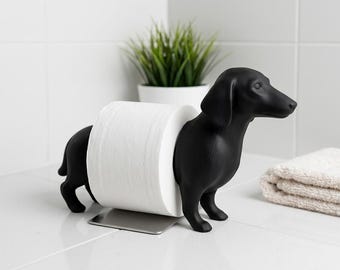 May include: A matte black toilet paper holder shaped like a dachshund dog. The dog figure is split into two parts, with the front and back sections connected by a central metal base that supports a single roll of white toilet paper.