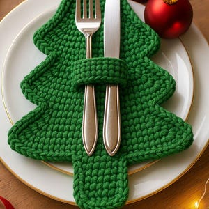 May include: A green, crocheted Christmas tree-shaped napkin holder with a fork and knife. The holder sits on a white plate with a gold rim, next to red ornaments. Festive holiday table setting.