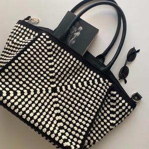 May include: Black and white crocheted tote bag with black leather handles and trim. The bag contains a black book and a pair of black sunglasses. The bag has a zip closure and a gold-coloured zip pull.