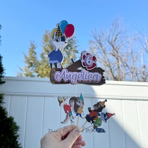 May include: A cake topper set featuring characters from the animated series "The Backyardigans". The set includes a log with the name "Angelica" and the number "6" on it, a raccoon, a squirrel, and a skunk. The characters are all wearing party hats.