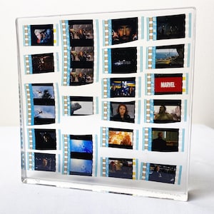 May include: A clear acrylic block containing multiple vertical strips of 35mm film featuring scenes from a Marvel movie. The film frames display various characters, action sequences, and the red Marvel logo, all preserved within the transparent material.