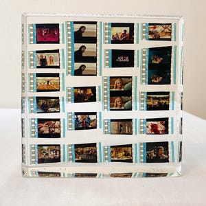 Könnte beinhalten: Ein klarer, rechteckiger Block mit Filmstreifen mit Filmszenen. Die Filmstreifen sind in einem Raster angeordnet, mit blauen Klebebandakzenten. Der Block wird auf einer weißen Oberfläche präsentiert.