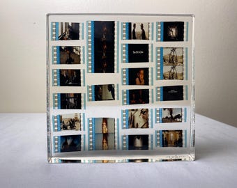 Op de afbeelding: Een helder acrylblok met filmstrips met filmscènes. De filmstrips hebben een blauw-witte rand. Sommige strips tonen tekst, waaronder "Once Upon a Time in the West". Het blok staat op een wit oppervlak.