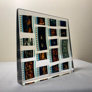 May include: A clear, rectangular acrylic block displaying film strips with various movie scenes. The film strips have a white background with teal and gold borders. The block is on a white surface.