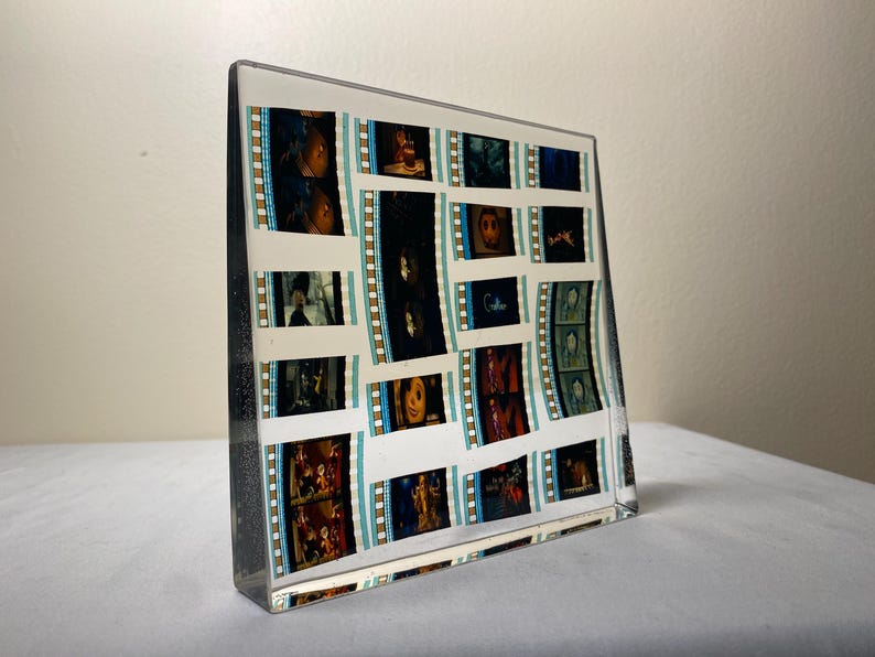 May include: A clear, rectangular acrylic block displaying film strips with various movie scenes. The film strips have a white background with teal and gold borders. The block is on a white surface.