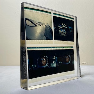 May include: A rectangular clear acrylic block containing three embedded 35mm film strips. The top two frames show a dark metallic surface and a figure, while the bottom frame displays a wide shot of a person in a space helmet against a dark background.