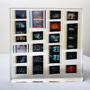 May include: A clear, square acrylic block containing multiple strips of 35mm film. The film frames display various scenes, including a map, a title card, and character portraits. The film strips are arranged in a grid pattern within the transparent material.