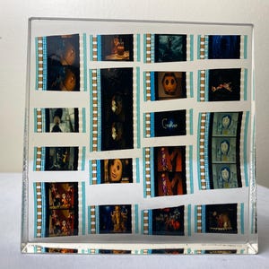 May include: A clear acrylic block featuring film strips with scenes from the movie "Coraline." The film strips are arranged in a grid pattern, with the word "Coraline" visible. The film strips have a blue and white border.