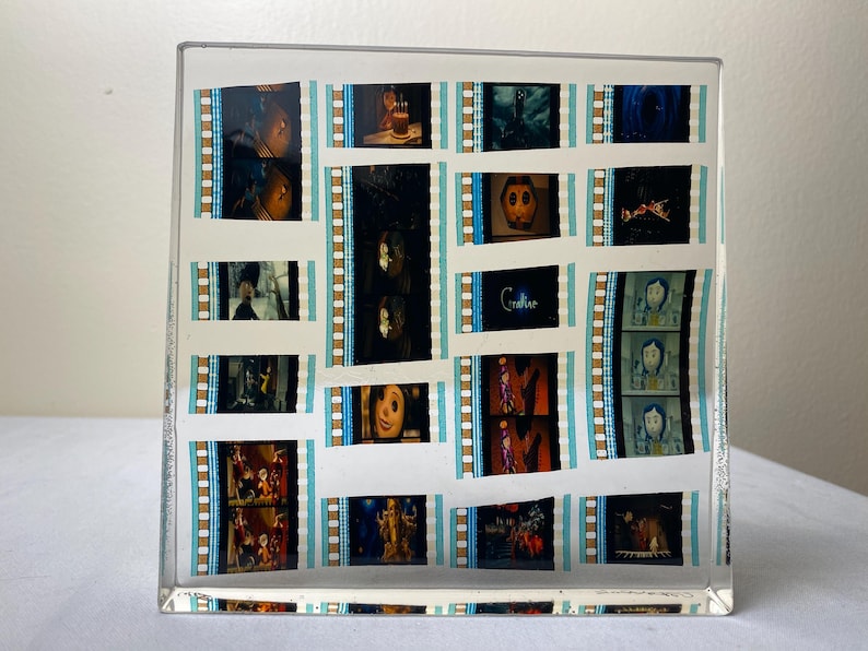 May include: A clear acrylic block featuring film strips with scenes from the movie "Coraline." The film strips are arranged in a grid pattern, with the word "Coraline" visible. The film strips have a blue and white border.