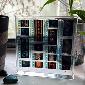 May include: A clear, rectangular resin block encasing film strips with various movie stills. The film strips have a teal and white border. The images within the film strips are in color and black and white. The block is displayed on a white surface.