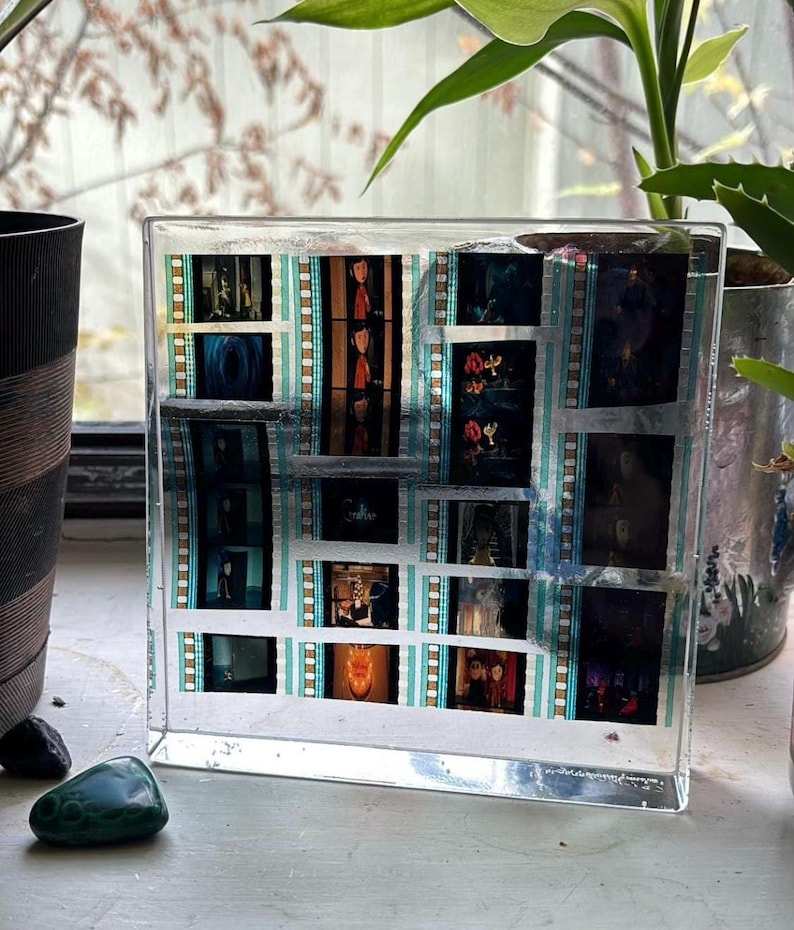 May include: A clear, rectangular resin block encasing film strips with various movie stills. The film strips have a teal and white border. The images within the film strips are in color and black and white. The block is displayed on a white surface.